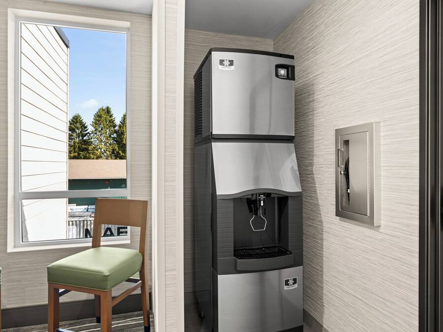 A compact kitchenette with a built-in refrigerator, a water dispenser, and a microwave. There is a window providing a view of trees and a building outside. Two chairs are placed in front of the window.