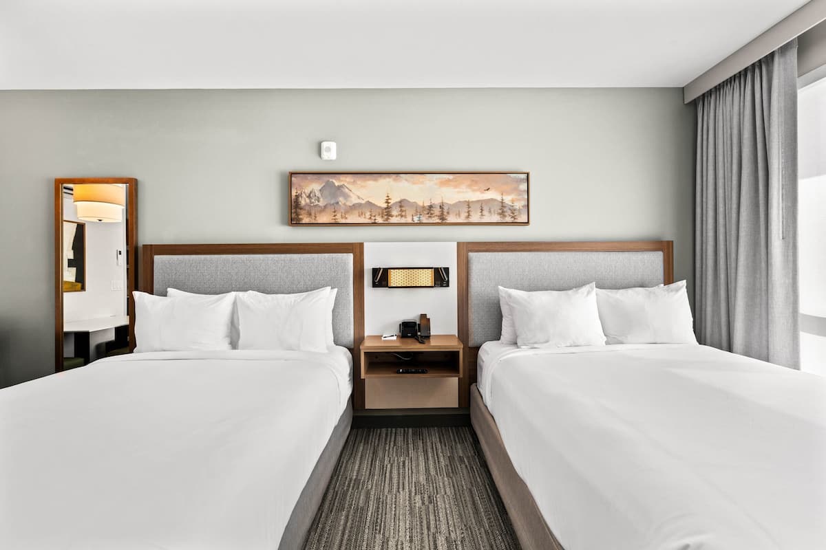 Ein Hotelzimmer mit zwei Betten, einem Nachttisch mit Lampe, einem Bild an der Wand und einem Fenster mit Vorhängen.