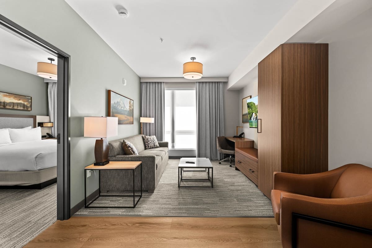 Ein modernes Hotelzimmer mit Bett, Sofa, Ohrensessel, Schreibtisch und einem großen Fenster mit Vorhängen.