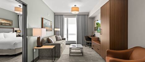 A modern hotel room with a bed, sofa, armchair, desk, and a large window with curtains.
