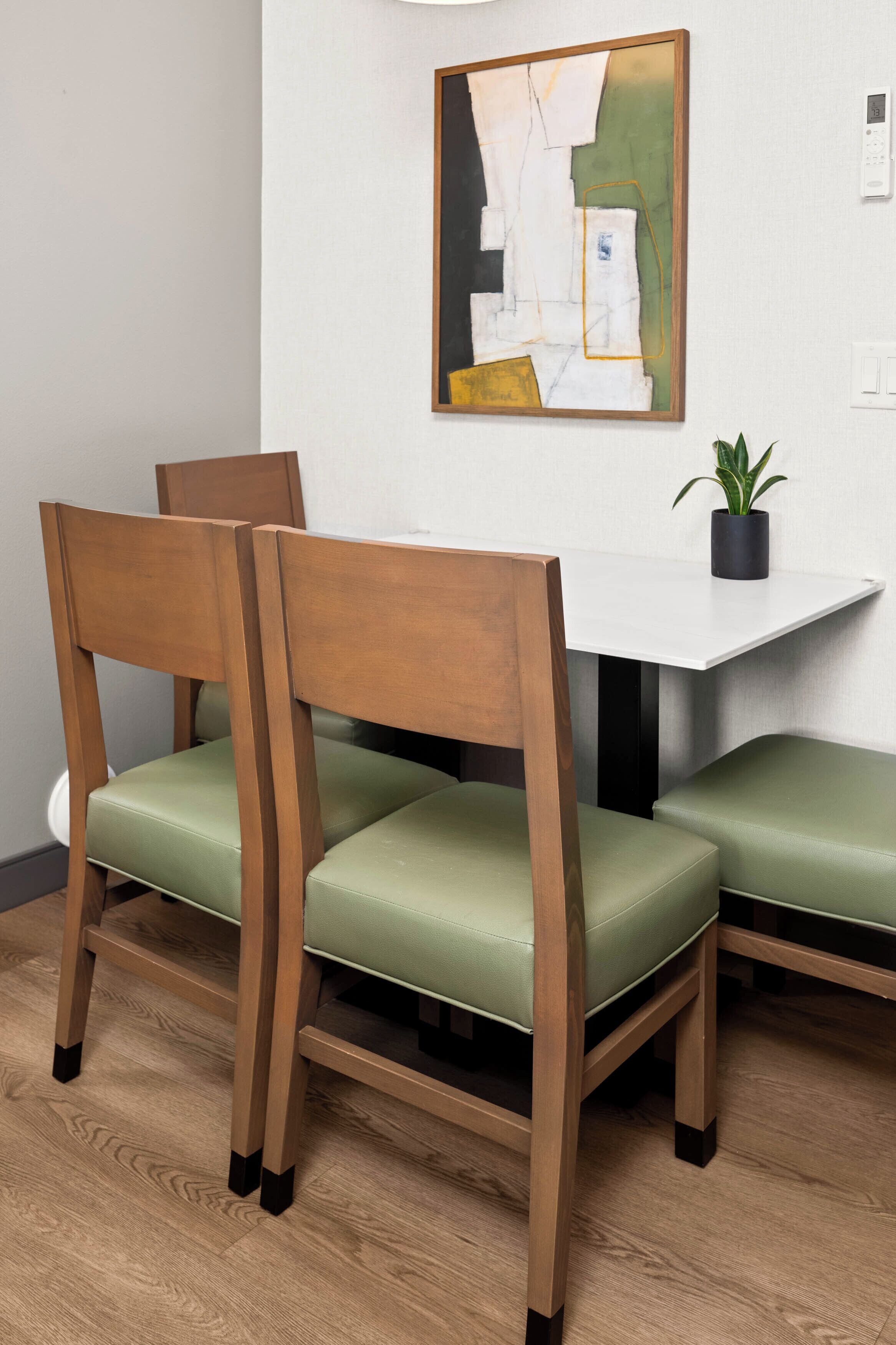 Een eethoek met een witte tafel, groene stoelen met kussens en een modern abstract schilderij aan de muur.