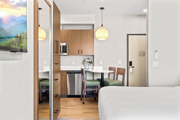 A modern hotel room with a bed, a dining area with a table and chairs, a kitchen with a microwave, and a view of a mountain landscape.