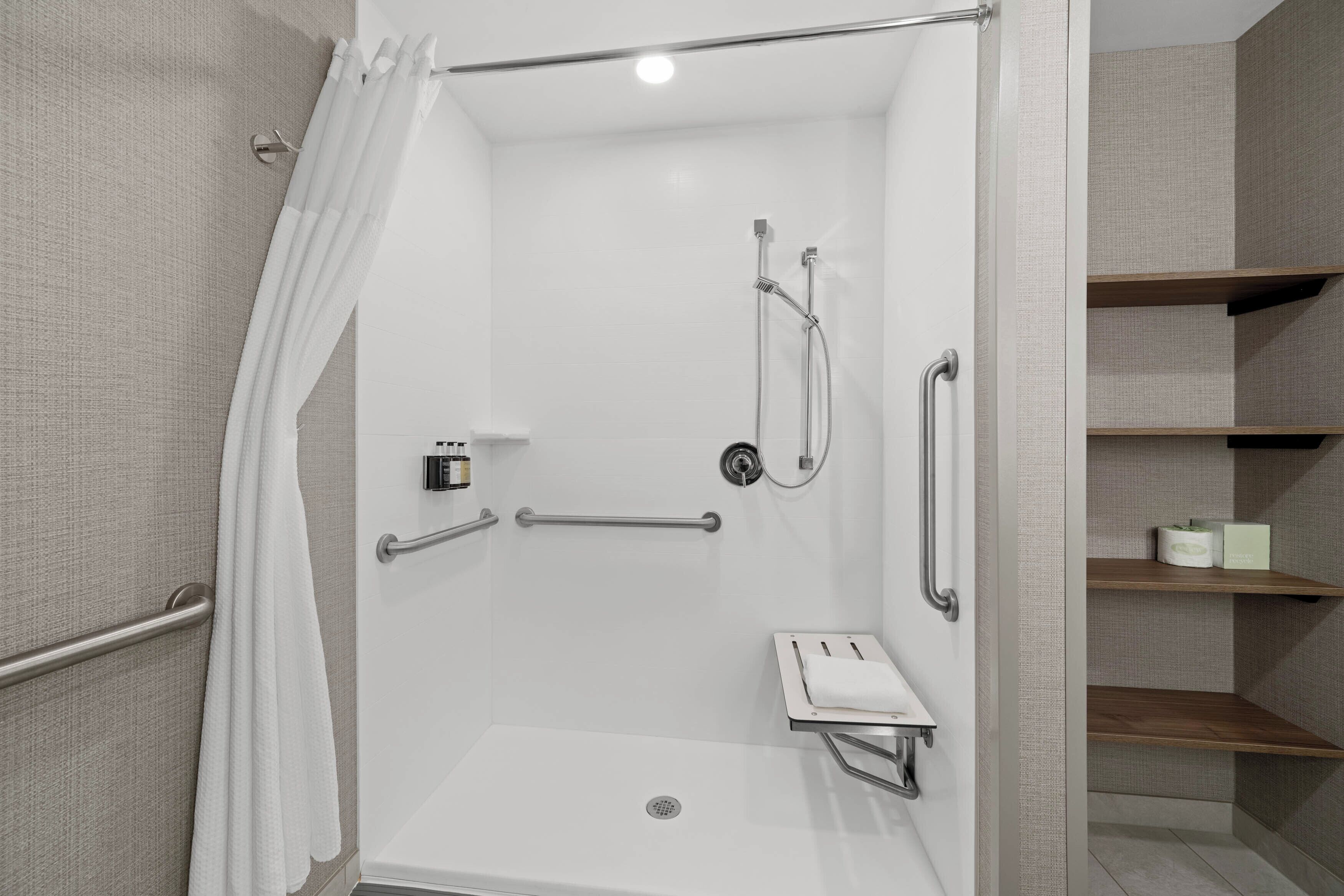 A modern shower area with a glass door, grab bars, a handheld showerhead, and a small shelf with toiletries.