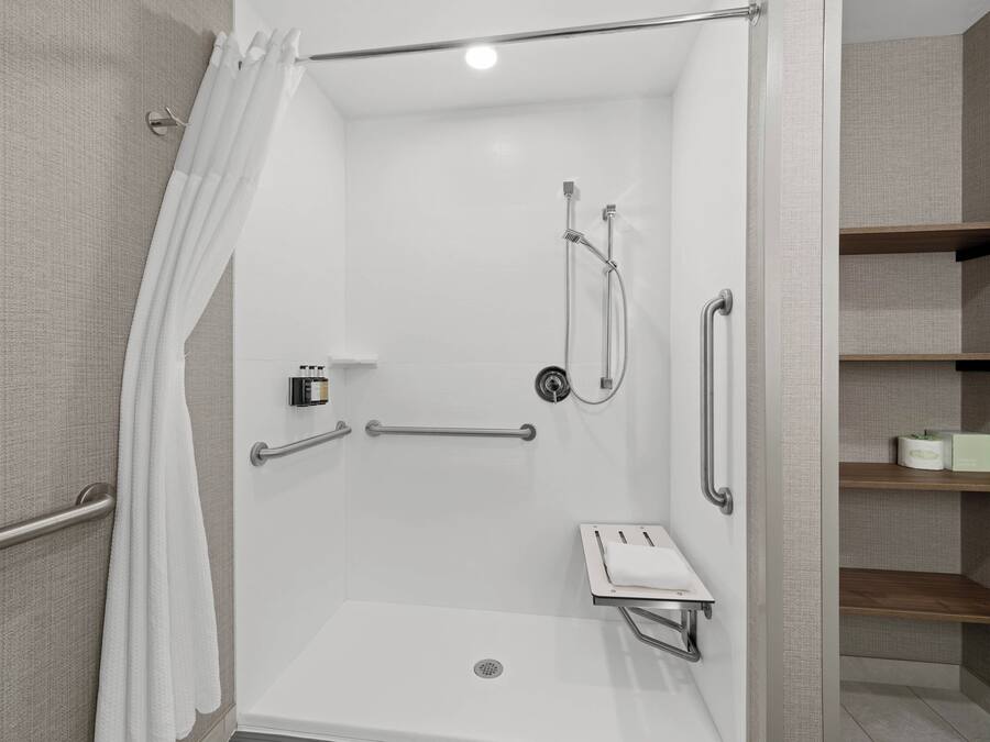 A modern shower area with a glass door, grab bars, a handheld showerhead, and a small shelf with toiletries.