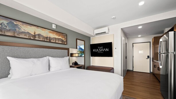 A hotel room with a large bed, a painting on the wall, a TV, and a kitchen area with a refrigerator. - The Kulshan Hotel (Sedro-Woolley)
