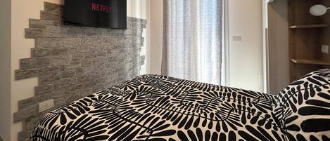 A bedroom with a stone wall, a bed with a patterned bedspread, a flat-screen TV mounted on the wall, and a window with curtains.