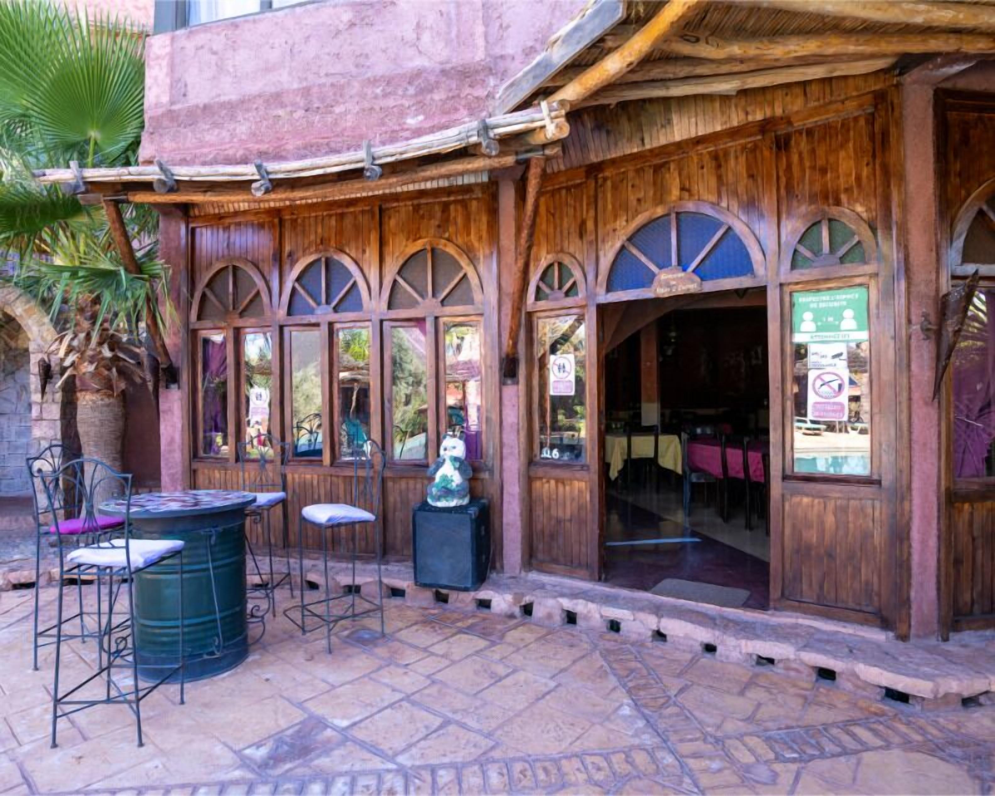 An outdoor seating area with wooden tables and chairs, a stone floor, and a palm tree.
