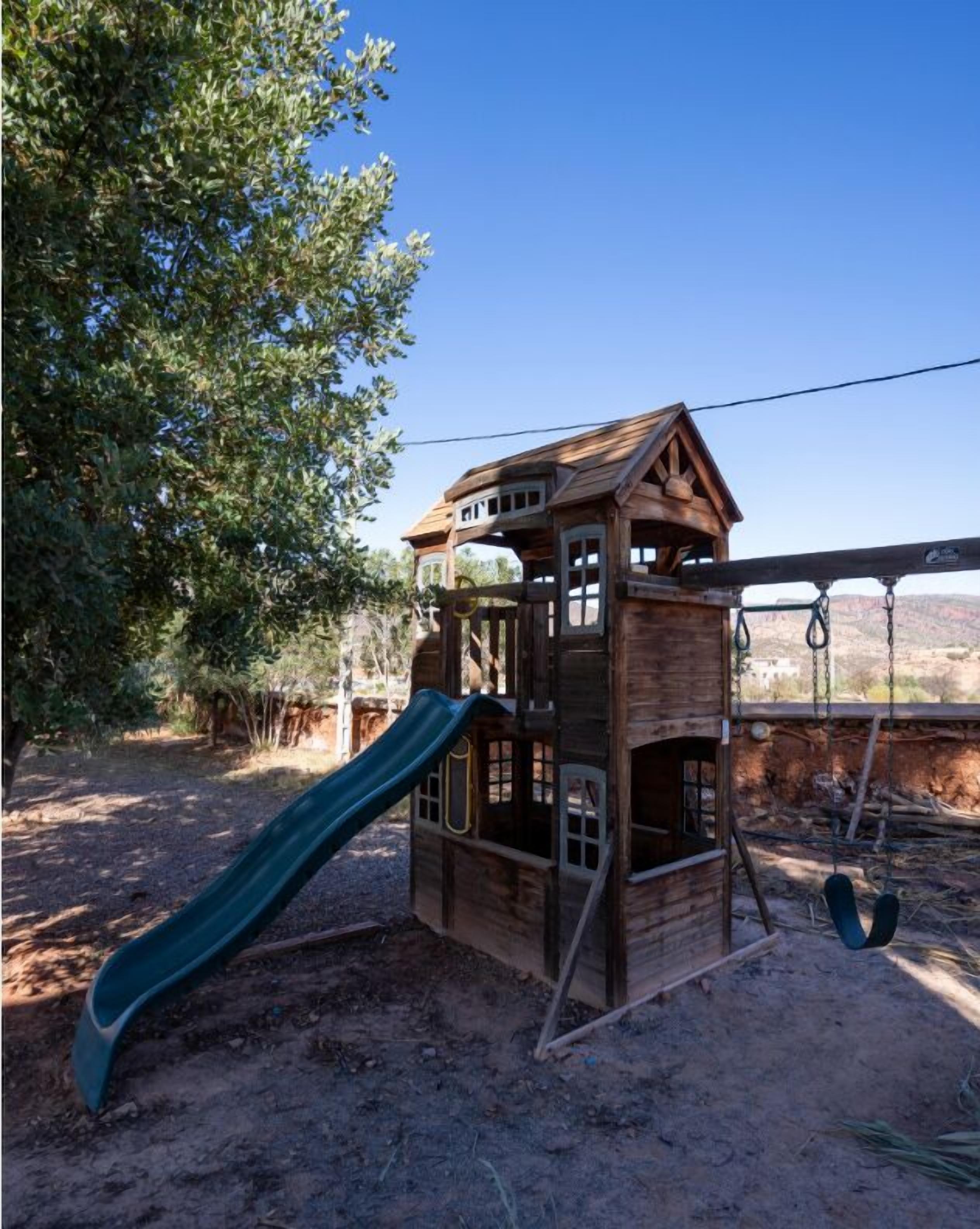 A wooden playhouse with a slide and swing set, surrounded by trees and a desert landscape.