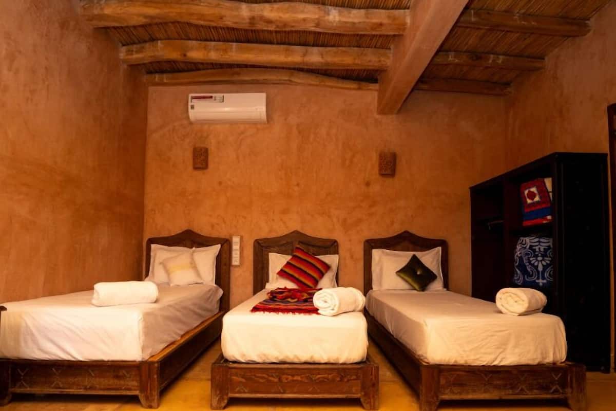 A room with three wooden beds, each with white linens and pillows, a wooden wardrobe, and a ceiling with exposed wooden beams.