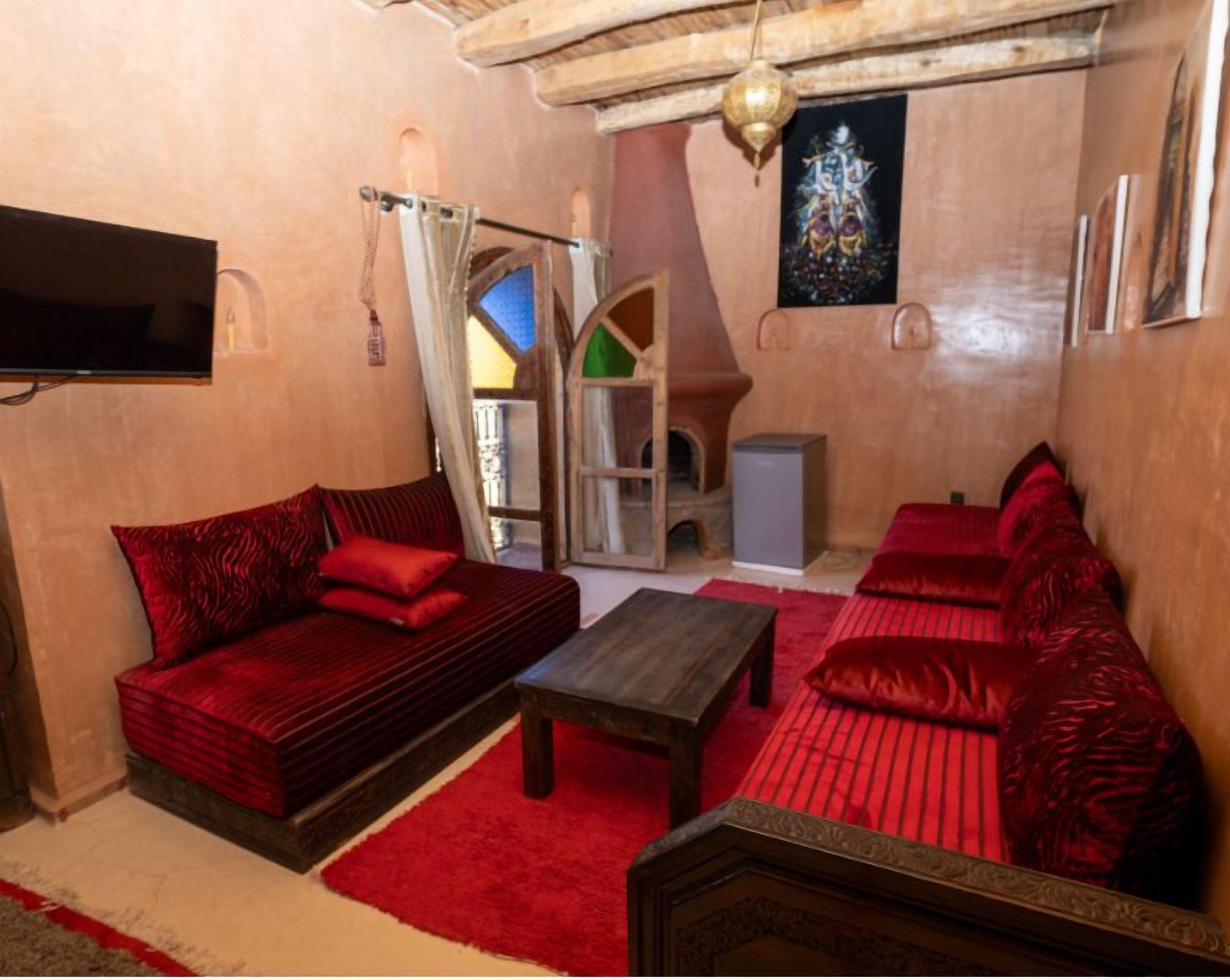A cozy room with a red sofa, a wooden coffee table, and a red cushioned bench. A television is mounted on the wall, and a decorative item hangs above the seating area.