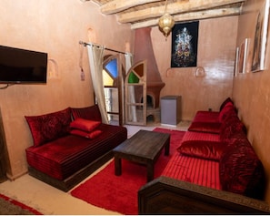 A cozy room with a red sofa, a wooden coffee table, and a red cushioned bench. A television is mounted on the wall, and a decorative item hangs above the seating area. - Palais d'Ouzoud (Ait Taguella)