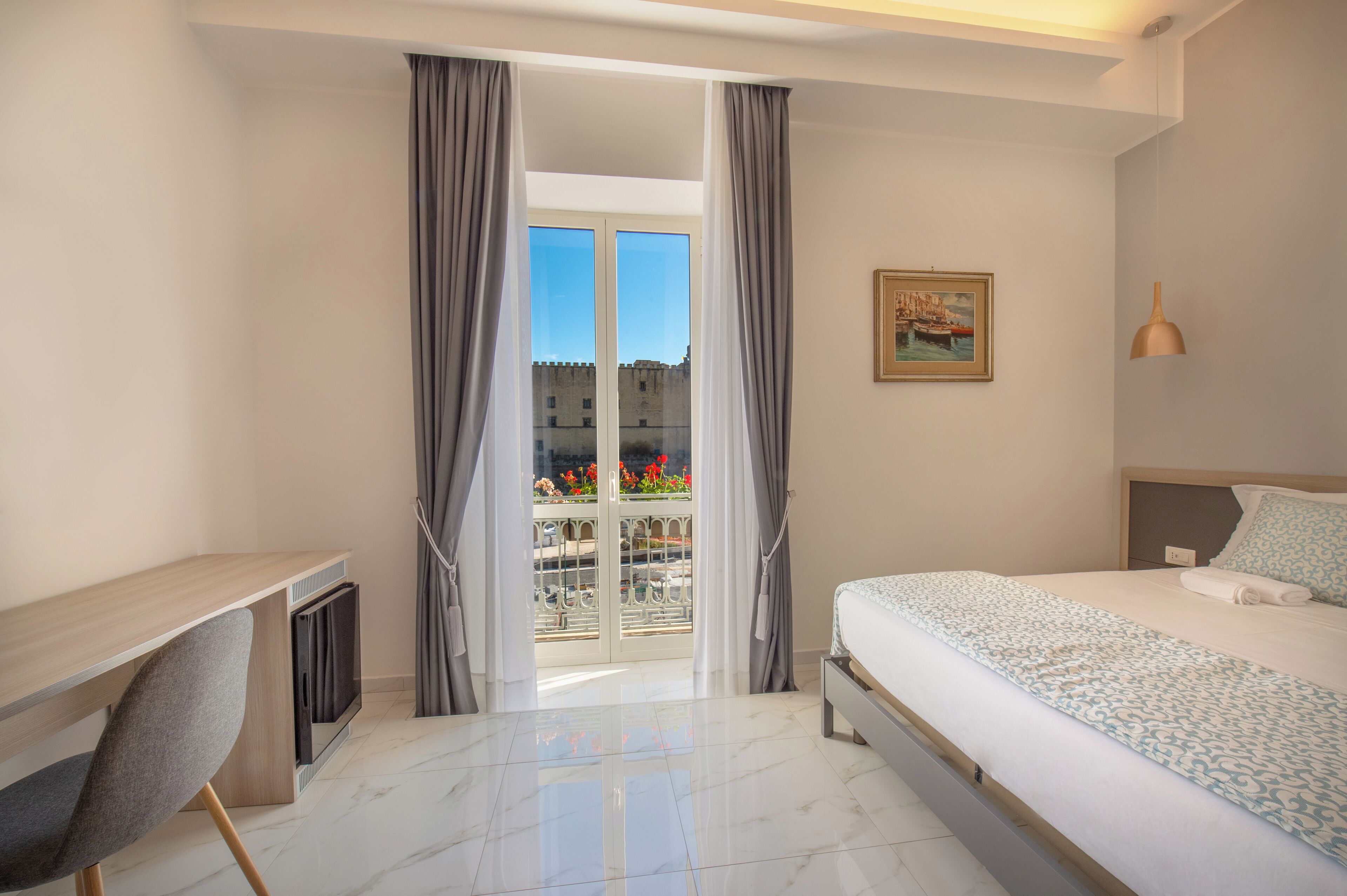 A hotel room with a bed, a desk, and a balcony with a view of a building and flowers.