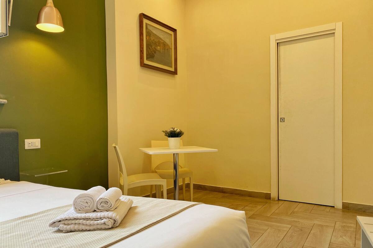 A hotel room with a bed, a small table with a plant, a chair, and a door.