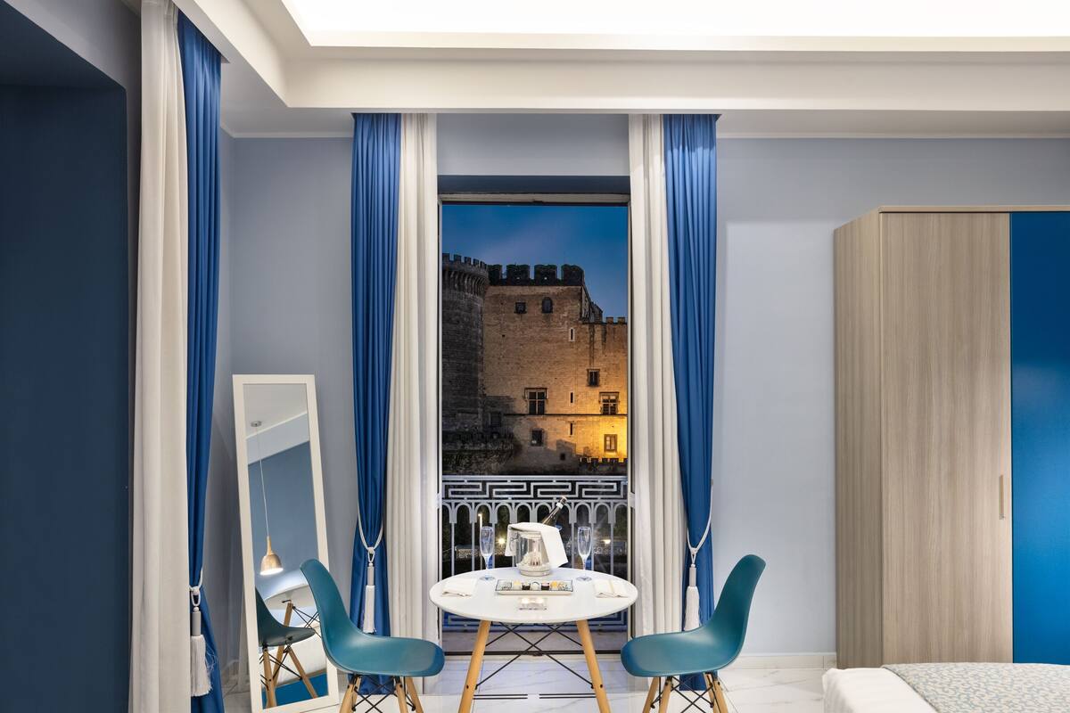 A modern room with a balcony, a round table with chairs, a wardrobe, and a view of a historic building.