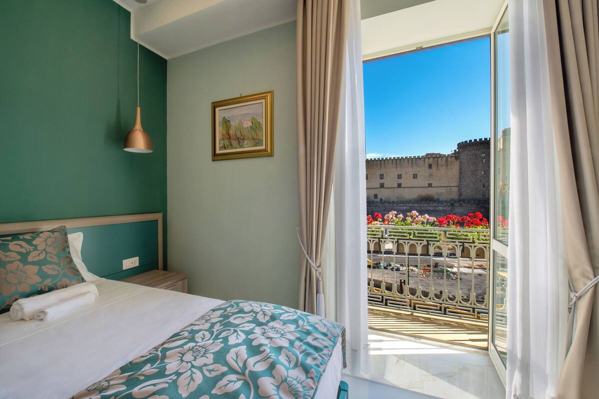 A bedroom with a bed, a nightstand, a hanging lamp, a window with curtains, and a view of a castle through the window.
