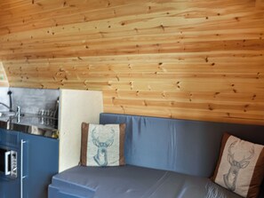 Una habitación con techo de madera, un sofá azul y una zona de cocina con refrigerador y lavamanos.
