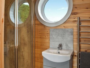 Un baño con cabina de ducha de vidrio, un ventanal redondo, un lavabo con salpicadero gris y una pared de madera.