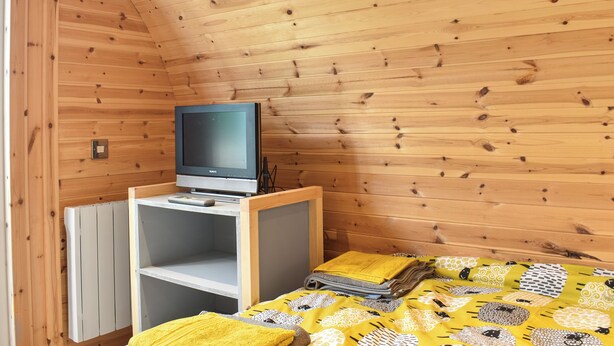 A small, cozy room with wooden walls and a sloped ceiling. A television is placed on a wooden stand against the wall, and a bed with a yellow patterned blanket is visible.