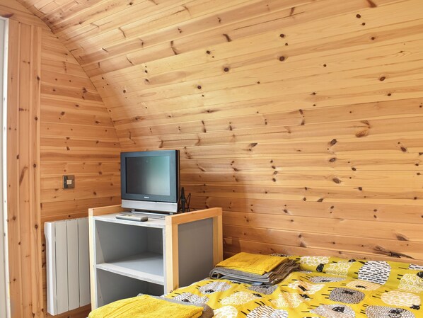 Una habitación pequeña y acogedora con paredes de madera y techo inclinado. Un televisor está sobre un mueble de madera adosado a la pared, y se ve una cama con un edredón amarillo estampado.