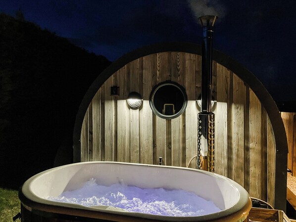 A hot tub in front of a wooden arched structure with a chimney emitting smoke.