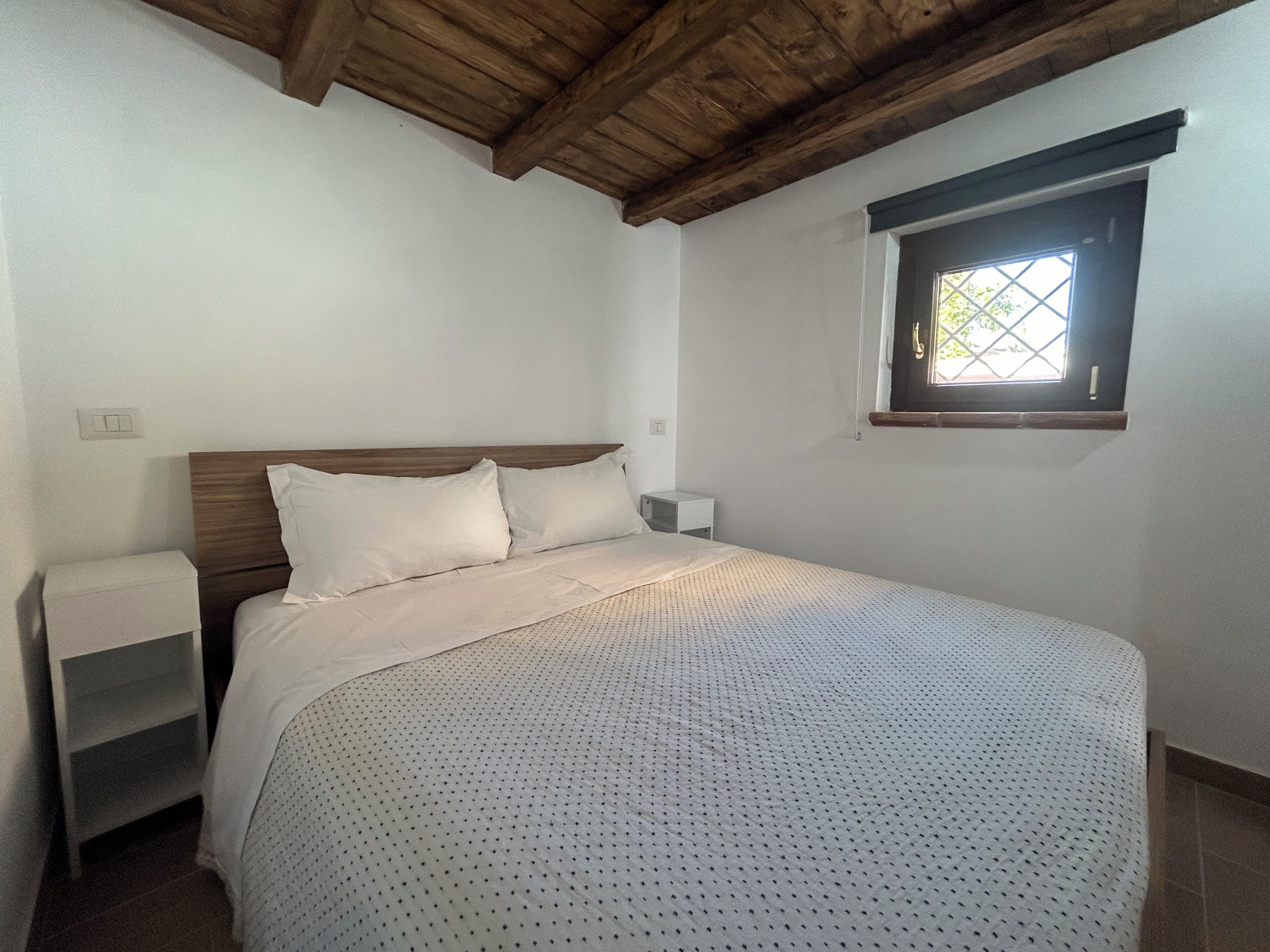 A bedroom with a wooden ceiling, a single bed with white bedding, a nightstand, and a window with a view of greenery.
