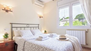 A bedroom with a bed, bedside lamps, a window with curtains, a radiator, and a nightstand with a plant. - Casa Hermosa (L'Escala)