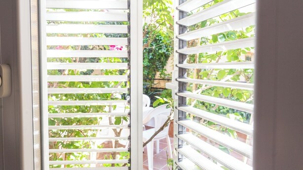 A view through white blinds with greenery outside, a white chair, and a stone wall.