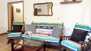 A living room with a sofa, chairs, a glass coffee table, a mirror, and a shelf with decorative items.
