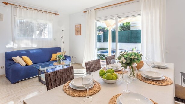 A modern living room with a blue sofa, dining table set with plates and glasses, and a view through large windows. - Casa Sunset (L'Escala)