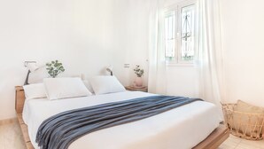 A neatly made bed with a blue blanket, a bedside table with a lamp, and a window with curtains. - Casa Sunset (L'Escala)