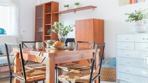 A dining area with a wooden table, wicker chairs, a cabinet with drawers, and a shelf with plants. - Casa Amapola (L'Escala)