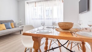 Un salon moderne avec une table à manger pour quatre personnes, un canapé avec des coussins, une grande fenêtre ornée de rideaux légers et un téléviseur à écran plat fixé au mur.
