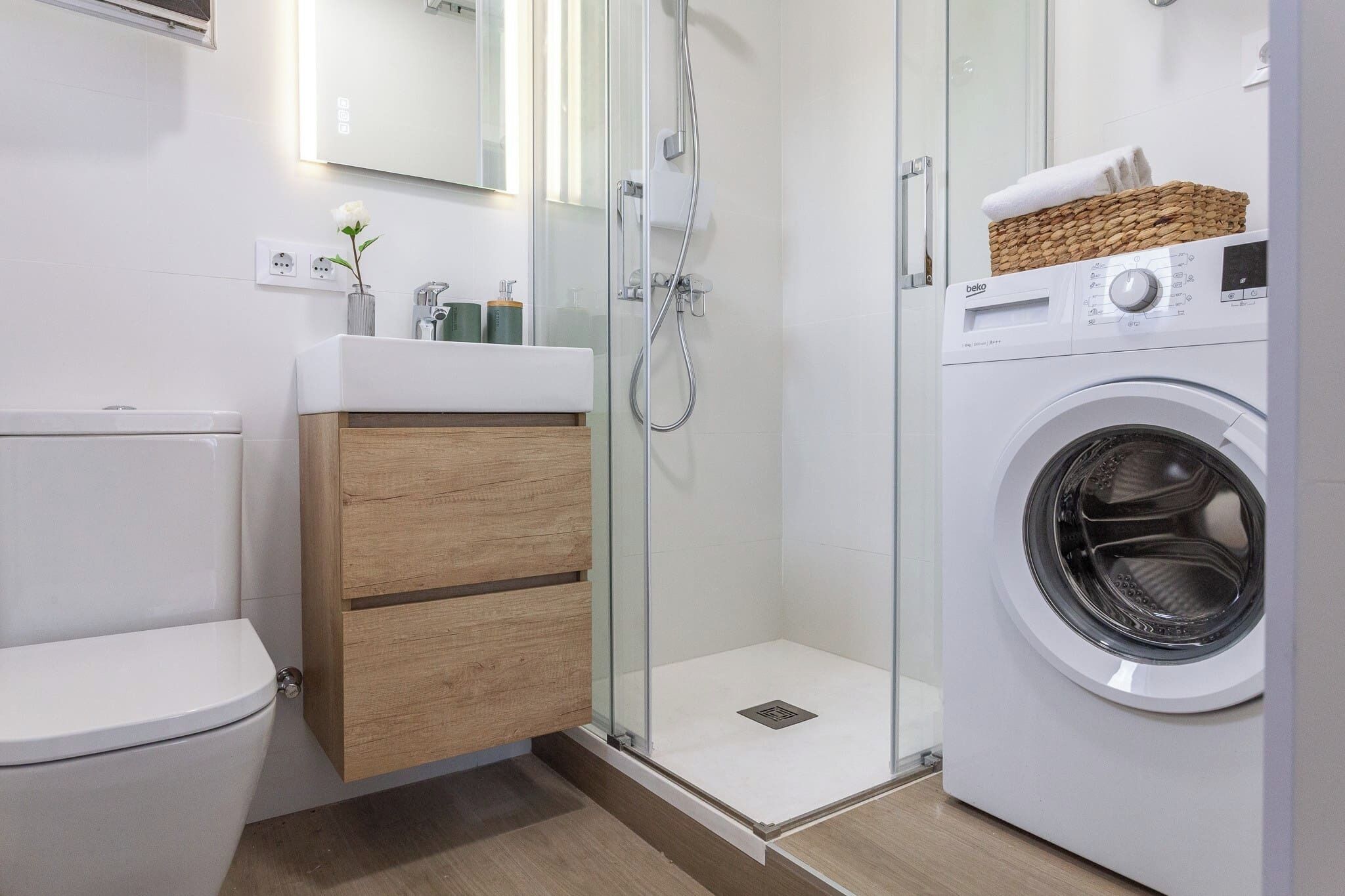 A modern bathroom with a toilet, a wooden vanity with drawers, a glass-enclosed shower, and a front-loading washing machine.