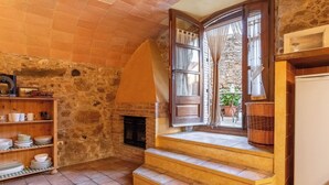 A rustic room with a stone wall, a fireplace, a wooden shelf with dishes, and a window with curtains. - Rural House Salvador XVIII (La Tallada d'Empordà)