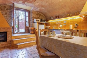 A cozy kitchen with a stone wall, a fireplace, a dining table set with cups and a cake stand, and a window overlooking a garden.