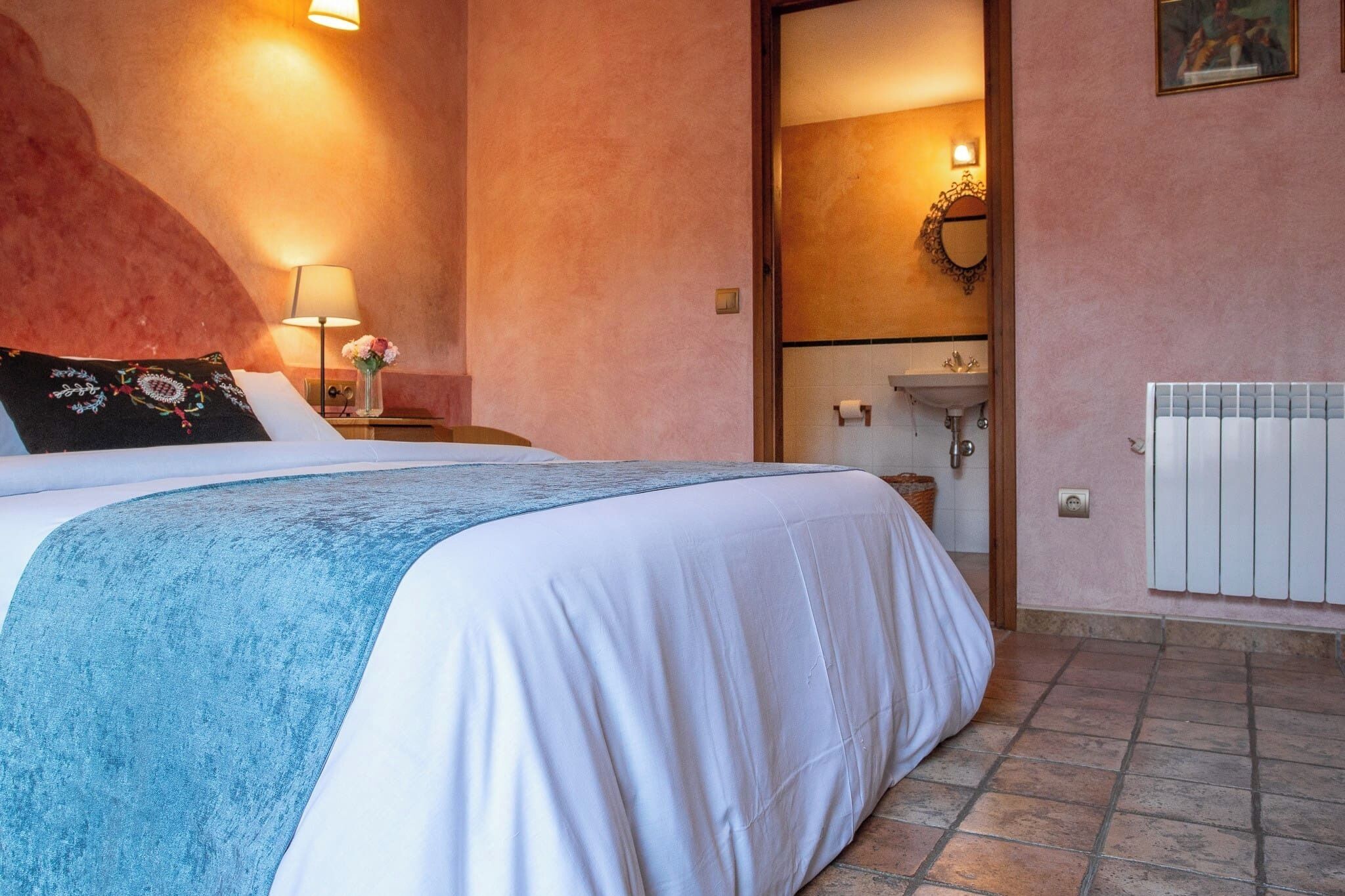 Un dormitorio con cama, mesita de noche, lámpara, estante en la pared y baño visible a través de una puerta abierta.