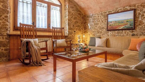 A cozy room with a stone wall, a wooden rocking chair, a sofa, and a table with glasses and a bowl of fruit.