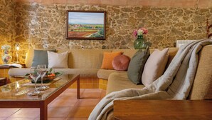 A cozy living room with a stone wall, a large painting, a sofa with cushions, a glass coffee table, and a vase with flowers.