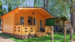 A wooden cabin with a porch and a small deck, surrounded by a grassy area and trees. - Bungalow Forest (L'Escala)