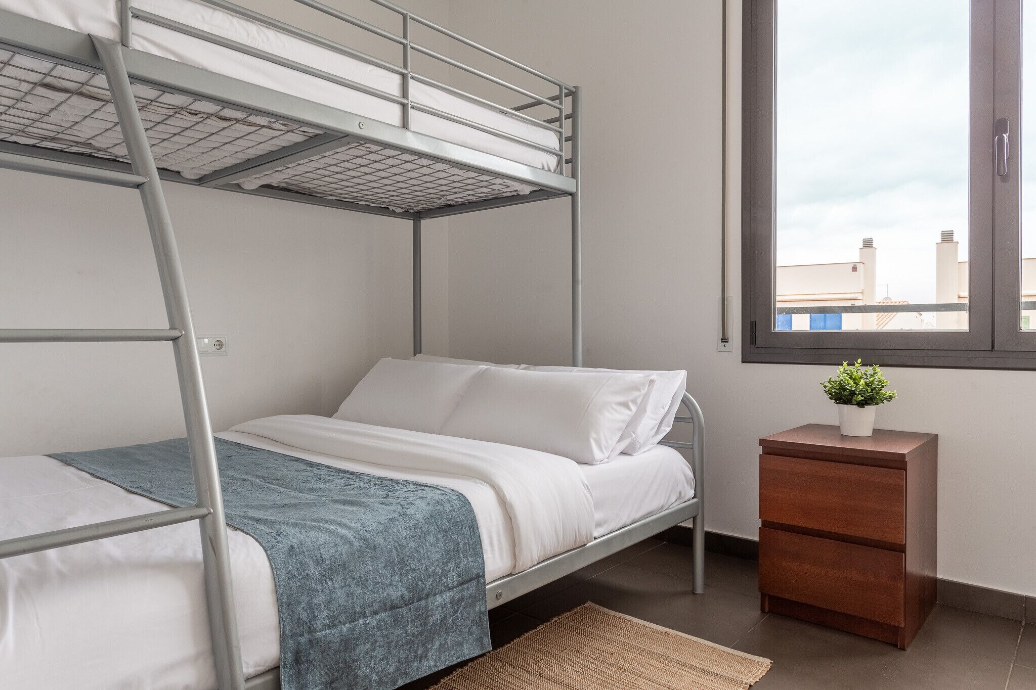 Una cama individual con estructura metálica, ropa de cama blanca y una manta azul. Hay una mesita de noche de madera con una planta encima y una ventana con vistas a edificios.