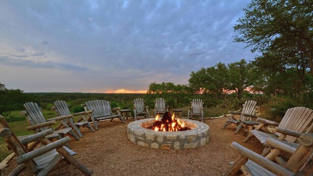 A circular stone fire pit surrounded by Adirondack chairs in a gravel area with a scenic view of trees and hills. - Cozy 1-bedroom cottage with outdoor fire pit (Kyle)