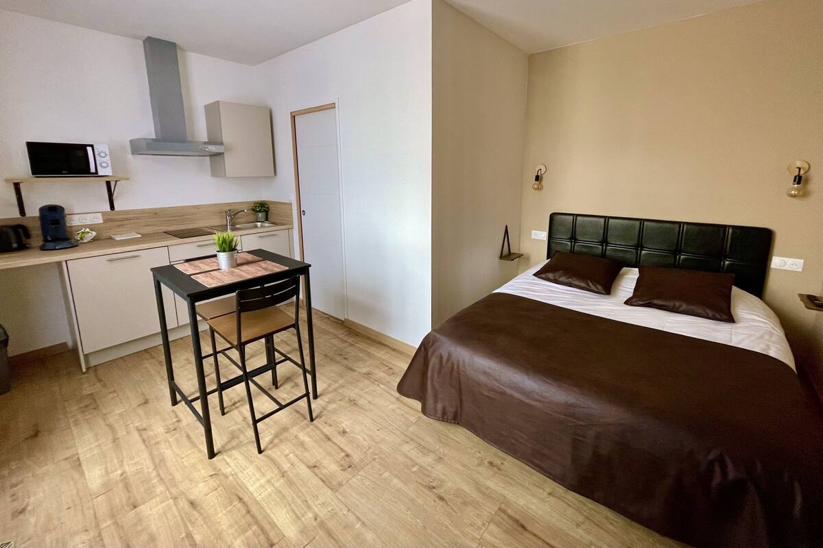 A compact hotel room with a kitchenette, a small dining table, and a bed with a dark headboard.