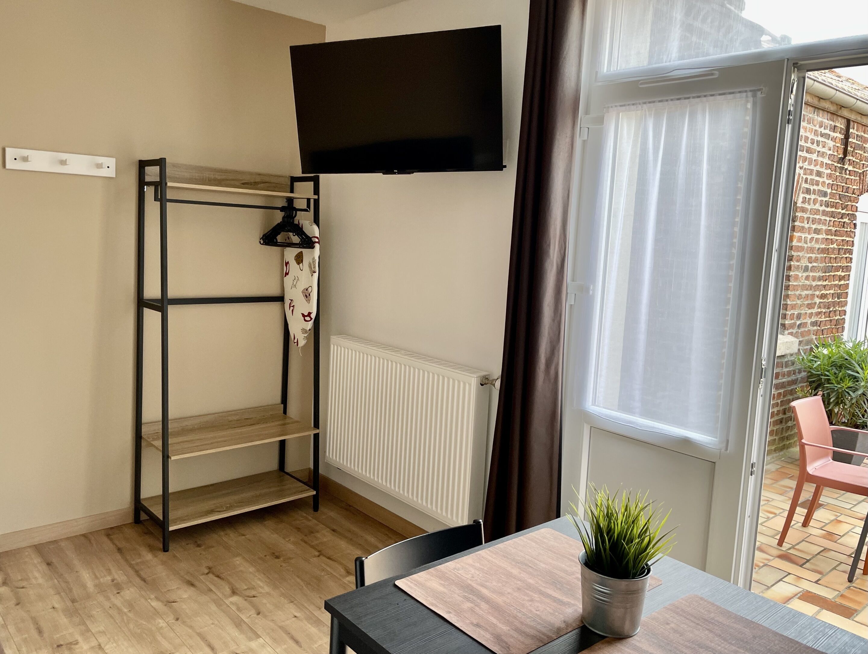Una habitación con una cama litera, un televisor de pantalla plana, un radiador, una mesa con una planta y vistas a un edificio de ladrillo a través de una ventana.