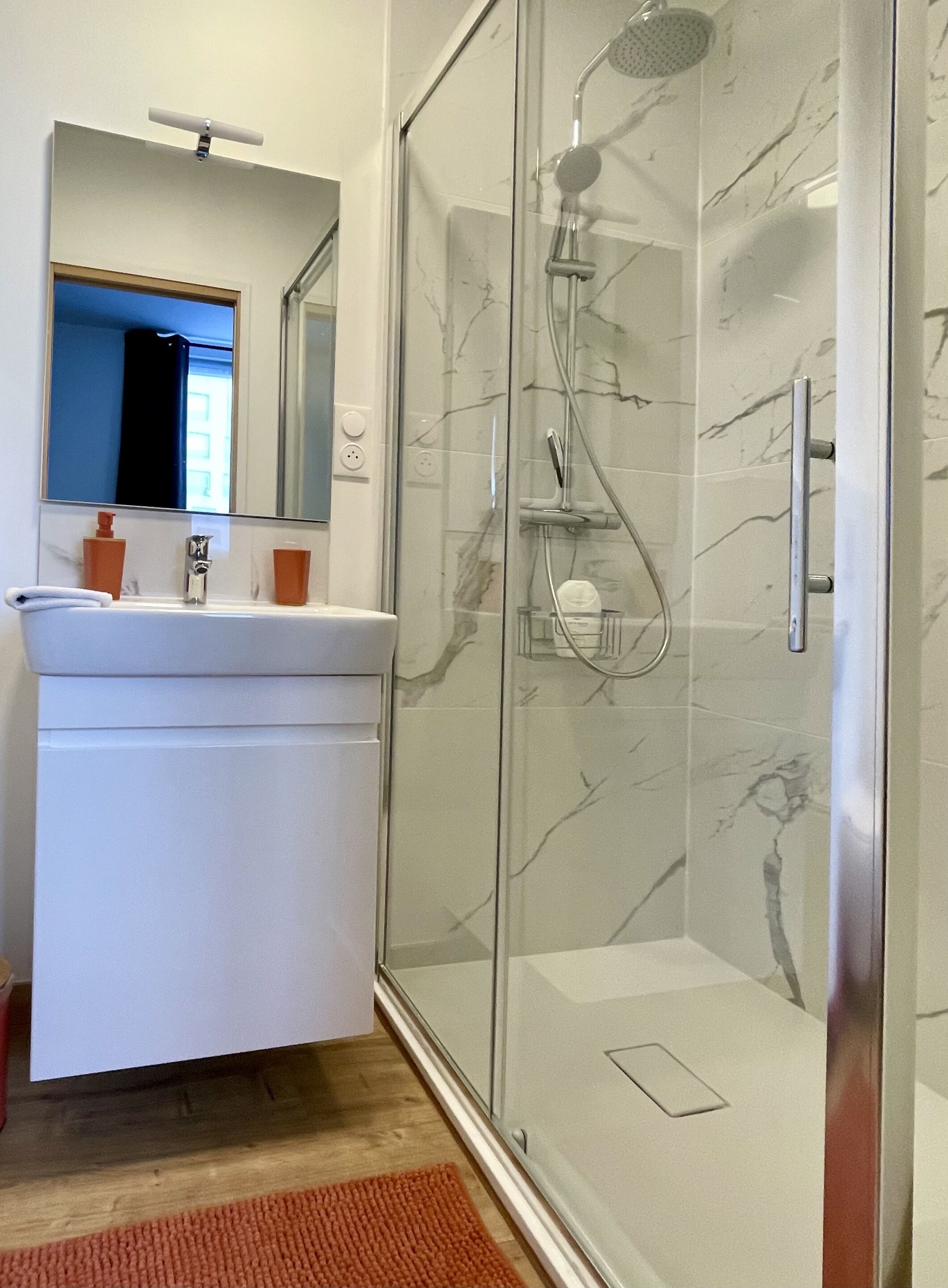 A modern bathroom with a glass-enclosed shower, a white sink with a mirror above it, and a wooden floor.
