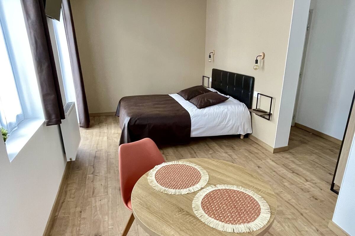 A hotel room with a bed, a round table with two placemats, a red chair, and a window with curtains.