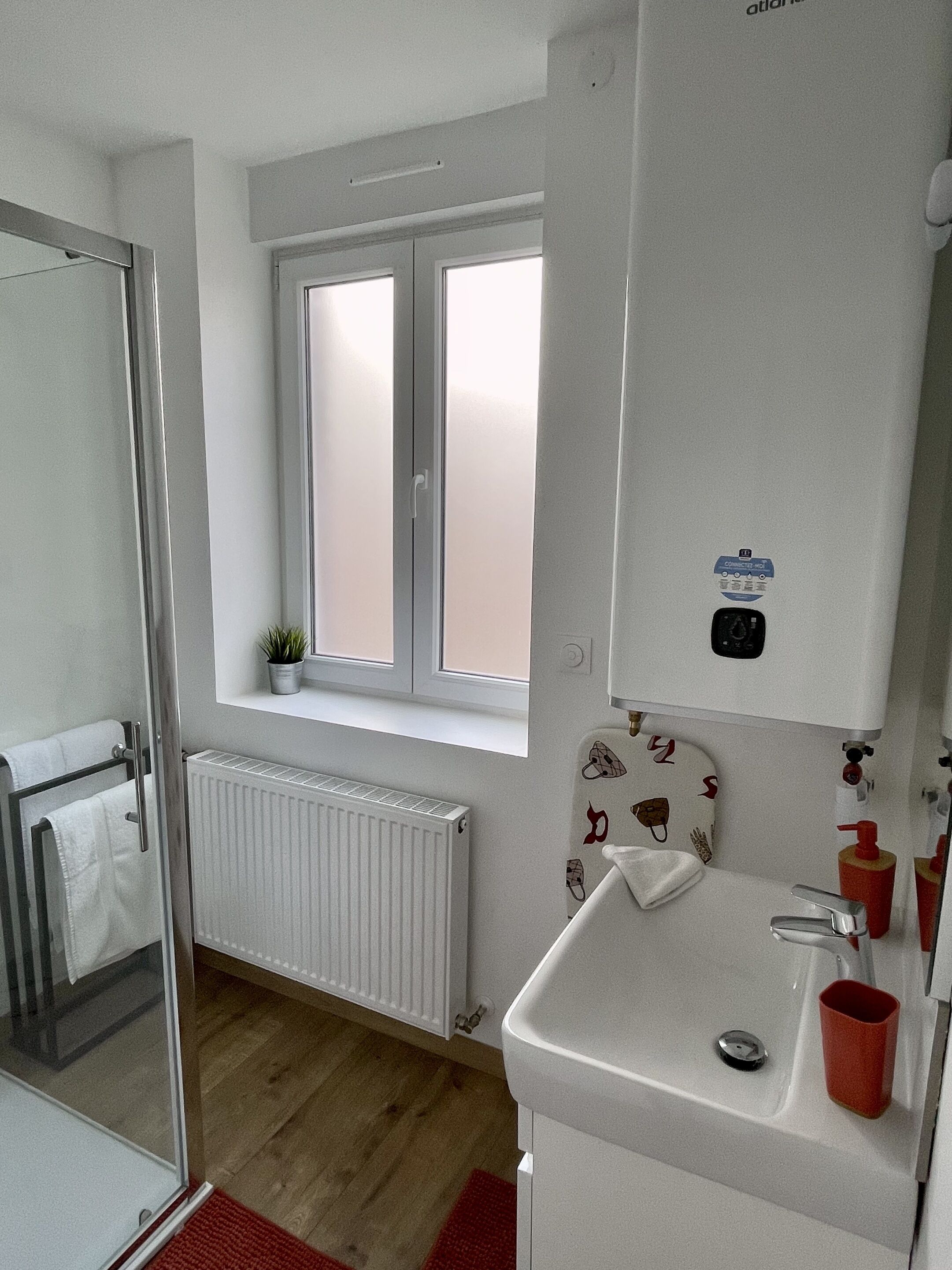 A modern bathroom with a glass shower door, a white sink with a red cup, a window with blinds, and a radiator.