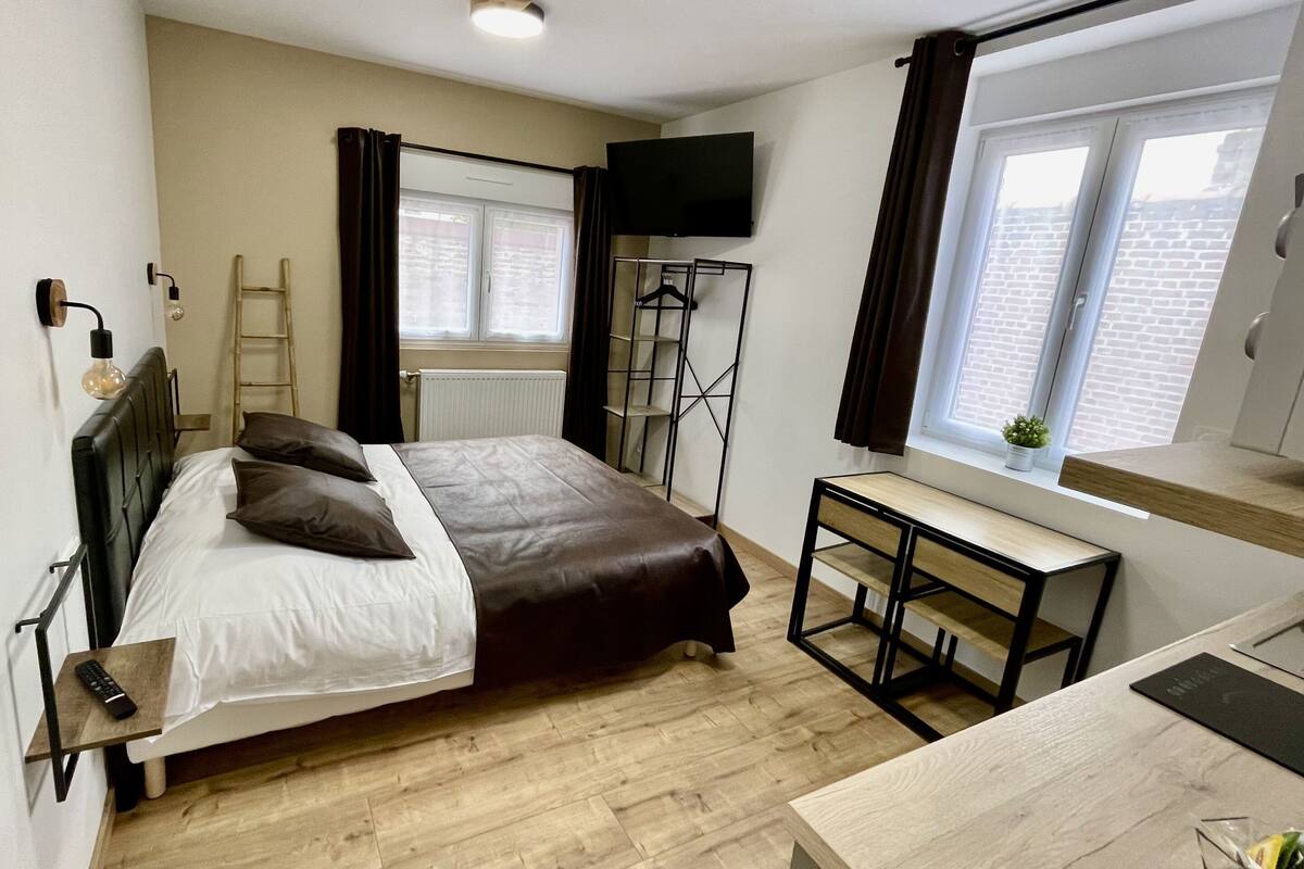 A bedroom with a bed, bedside table, and a desk with a chair. There is a window with curtains, a radiator, and a TV mounted on the wall.