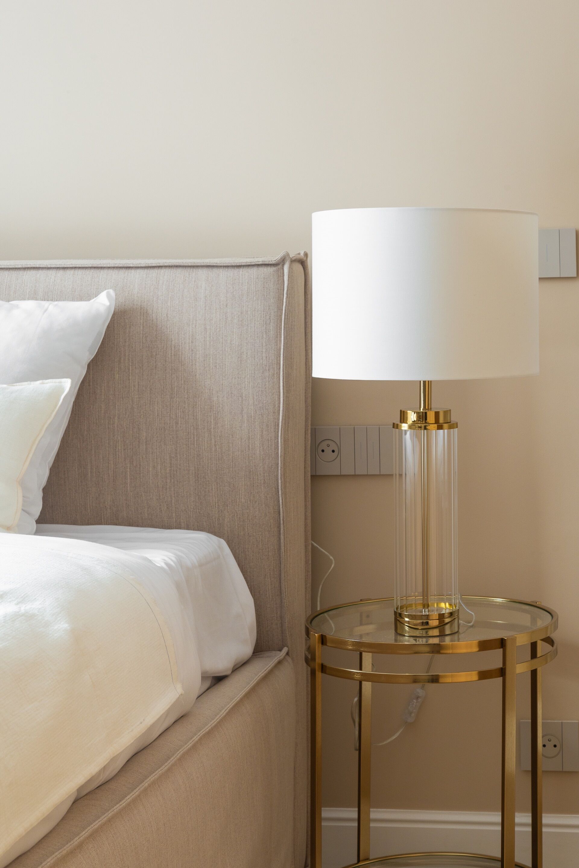 A bed with white bedding, a round gold-framed table lamp, and a wall socket.