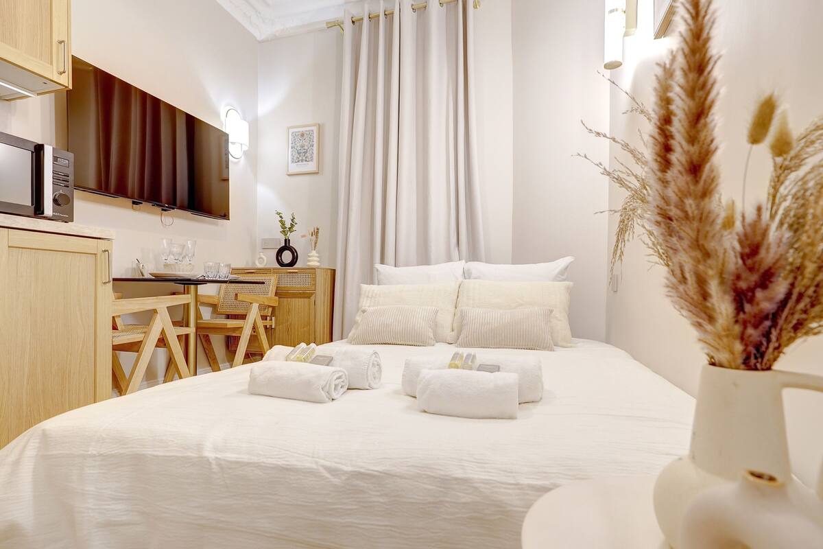 A hotel room with a bed, a desk, a chair, a TV, and a vase with dried plants.
