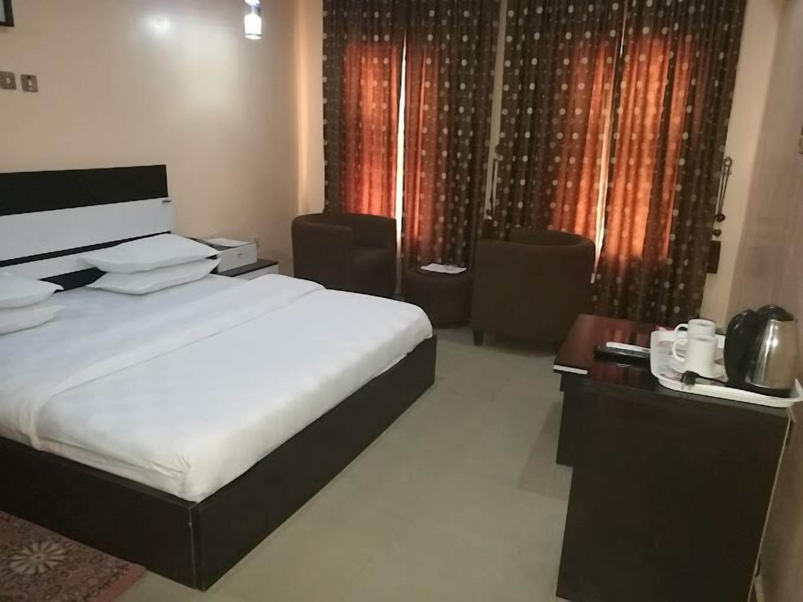 A hotel room with a bed, two chairs, a small table with a kettle and cups, and a window with curtains.
